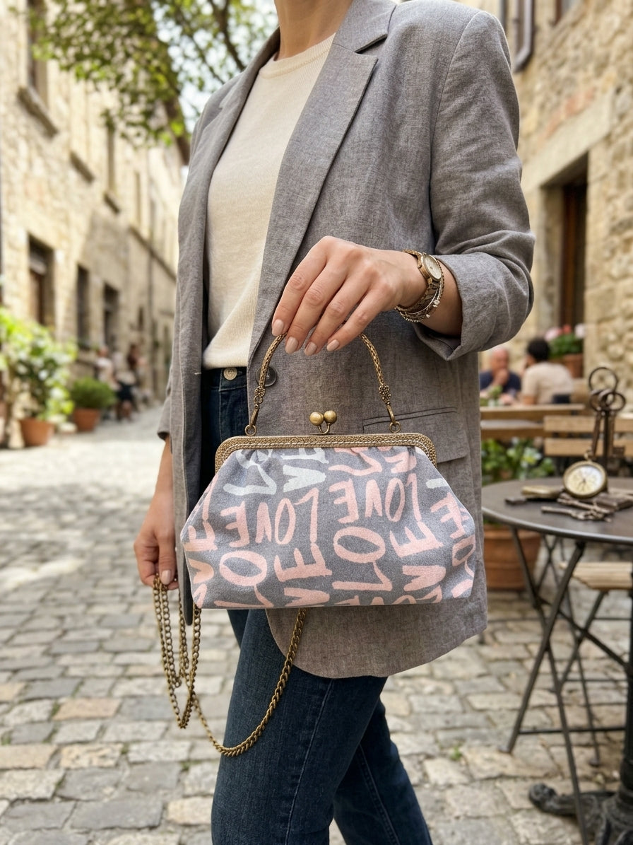 Graue Bügeltasche mit rosafarbenem Schriftmuster und antikgoldfarbener Kette als Schultertasche zu einem grauen Blazer und Jeans.